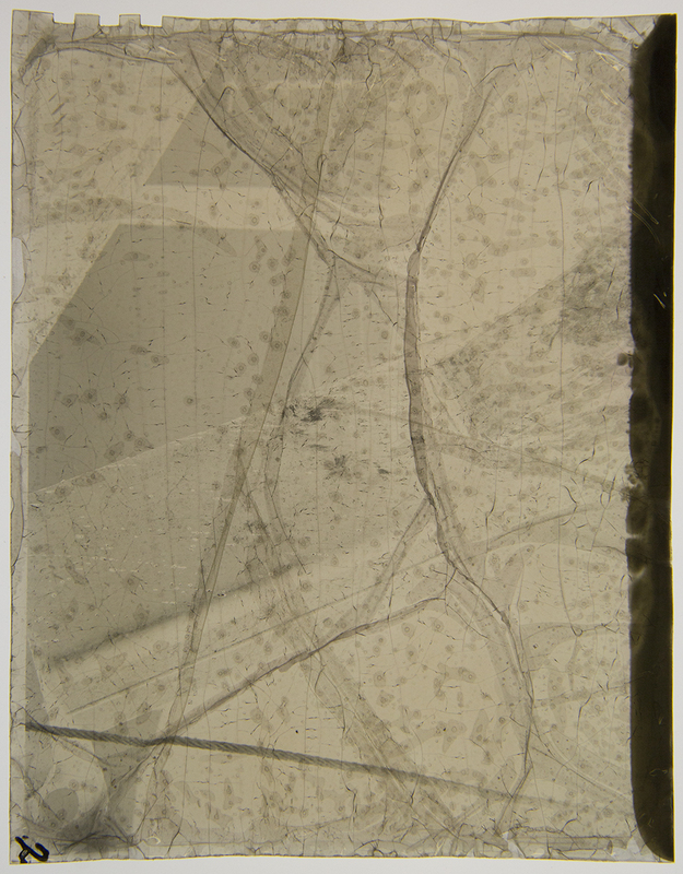 Imagen histórica en estado avanzado de degradación física que muestra fragmentos de una escena documental del periodo revolucionario mexicano. La descomposición química del soporte ha fracturado la emulsión, creando un entramado de grietas y pliegues que oscurecen los detalles originales de la composición, dejando apenas visibles formas que sugieren estructuras arquitectónicas o figuras humanas en un entorno urbano.