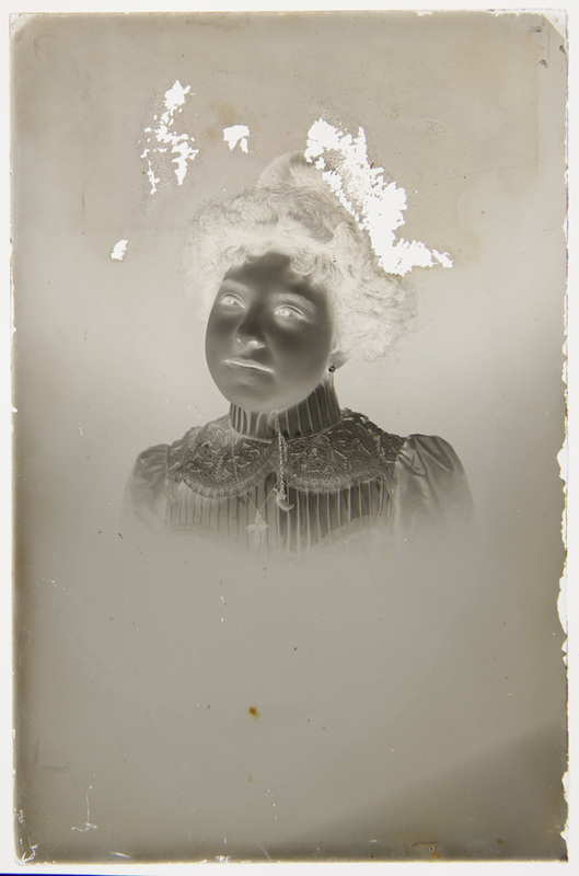 Retrato de estudio de una mujer joven con vestimenta formal de finales del siglo XIX o inicios del XX. La figura porta una blusa de cuello alto decorada con encaje intrincado y joyería que incluye una cruz y un dije de medialuna. Su peinado recogido y el estilo de la prenda sugieren una pertenencia a la clase media o alta urbana de la época.