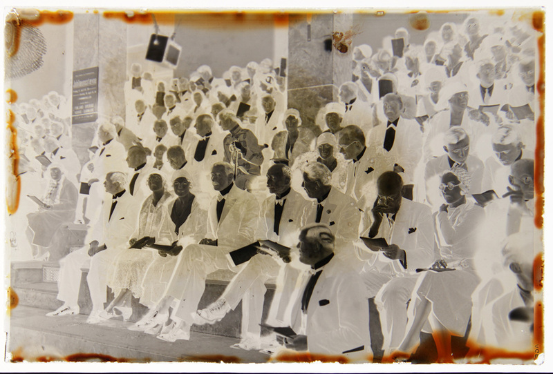 Imagen en negativo que captura a una multitud sentada en gradas durante una ceremonia oficial. Los asistentes, vestidos con ropa formal de gala, incluyen hombres de edad avanzada con trajes oscuros y mujeres con vestidos de media gala y sombreros. Un cartel informativo en una columna a la izquierda anuncia un evento conmemorativo relacionado con países escandinavos, sugiriendo una reunión diplomática o cultural internacional.