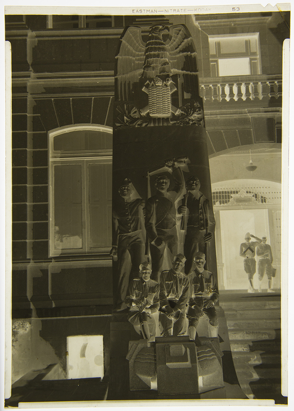 Fotografía en negativo que muestra un monumento conmemorativo de estilo modernista con relieves de cadetes y el escudo nacional mexicano en la parte superior. La escena se desarrolla frente a un edificio de arquitectura institucional o académica, donde se aprecia a dos individuos portando uniformes militares que caminan hacia un pórtico iluminado al fondo.