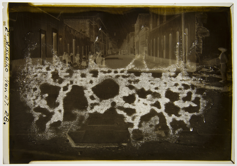 Escena urbana capturada en una calle de arquitectura colonial en México a finales de 1928. En la composición se aprecian diversas figuras humanas, incluyendo hombres con sombreros de ala ancha y mujeres con rebozos, situados frente a fachadas con puertas altas y molduras clásicas. La imagen documenta la atmósfera social y el entorno construido durante el periodo del Maximato.