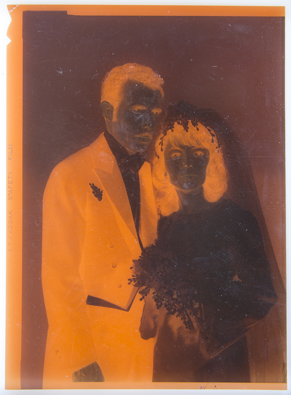 Un retrato nupcial de estudio que captura a una pareja joven en una pose formal. El hombre viste un esmoquin oscuro con pajarita y un boutonniere en la solapa, mientras que la mujer luce un vestido con detalles decorativos en el cuello y mangas, acompañado de un velo floral y un ramo de flores. La iluminación es controlada, destacando los rasgos de ambos sujetos en un encuadre de medio cuerpo.