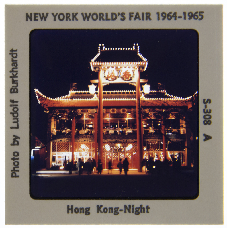 Vista aérea capturada durante la Feria Mundial de Nueva York de 1964-1965, centrada en el Pabellón de Nueva Jersey con su distintiva arquitectura de techos cuadrados y mástiles. En el plano medio destaca la Uniesfera rodeada de fuentes, mientras que al fondo se aprecia el Shea Stadium bajo un cielo despejado.
