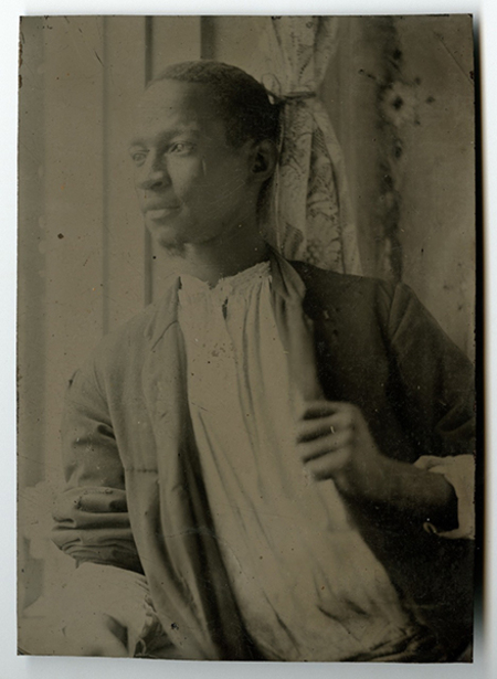 Retrato de busto de un hombre joven afrodescendiente mirando hacia un costado en pose reflexiva. Viste una camisa clara con cuello fruncido y una prenda exterior oscura abierta. El fondo muestra un interior sencillo con una cortina de motivos florales a la derecha, sugiriendo un entorno de estudio fotográfico de época.