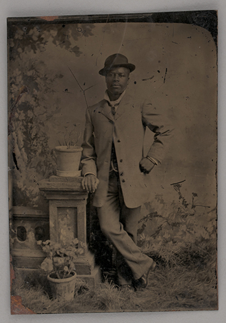 Un retrato de estudio de cuerpo completo que muestra a un hombre joven de ascendencia africana. El sujeto viste ropa formal del siglo XIX, incluyendo un sombrero hongo y un saco oscuro, posando con una mano en la cadera y apoyado en un pedestal con plantas frente a un telón de fondo pintado.