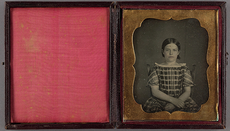 Retrato fotográfico de una niña sentada en una silla de madera, capturado a mediados del siglo XIX. La infante viste un vestido con patrón de cuadros, escote recto y mangas cortas abullonadas. La imagen destaca por su composición formal y frontal, típica de la época, presentada en un estuche protector con acabados en terciopelo rosado y marco de latón decorado.