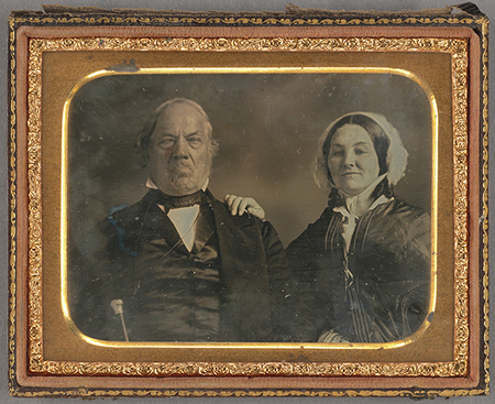 Retrato de estudio de mediados del siglo XIX de una pareja de edad avanzada en pose formal. El hombre, a la izquierda, viste un traje oscuro con un cuello rígido y corbatín, mostrando una expresión severa. A su lado, una mujer porta un vestido oscuro y una cofia blanca de encaje que enmarca su rostro, colocando su mano sobre el hombro del caballero en señal de unión familiar.