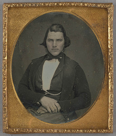 Retrato de medio cuerpo de un hombre joven con mirada directa y ojos claros. Viste indumentaria formal del siglo XIX, compuesta por una chaqueta oscura, camisa blanca de cuello alto y un lazo oscuro anudado al cuello. Se observa una leontina cruzando su chaleco y sus manos reposan tranquilas sobre su regazo, mostrando una pose solemne típica de la época.