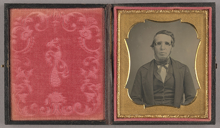 Retrato de estudio de medio cuerpo de un hombre adulto con patillas pronunciadas, vistiendo ropa formal de mediados del siglo XIX que incluye saco oscuro, chaleco y corbata. Destaca el uso de anteojos con cristales oscuros. La imagen está resguardada en un estuche abierto con una cubierta interior de terciopelo rojo decorada con un motivo de pavo real.