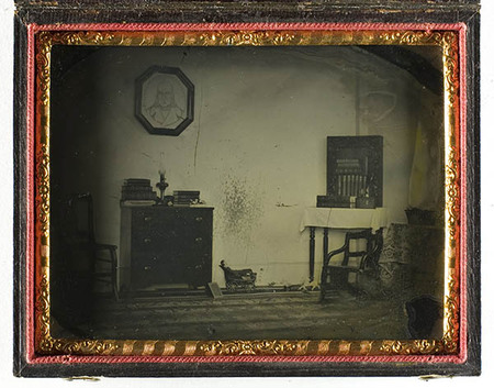 Vista interior de una habitación doméstica de mediados del siglo XIX. La escena incluye una cómoda con libros y una lámpara, un retrato octogonal colgado en la pared y una mesa con mantel blanco. En el suelo se aprecia un juguete pequeño, y a la derecha aparece una figura humana borrosa debido al movimiento durante la exposición prolongada.