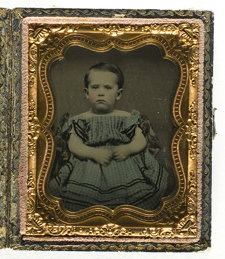 Retrato de estudio de un infante de corta edad, con cabello corto y expresión seria. Viste una prenda de patrón cuadriculado con cintas oscuras decorativas y hombros caídos, una moda infantil prevalente en el siglo XIX. El sujeto posa sentado en una composición de medio cuerpo, característica de la retratística comercial de la época.
