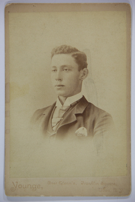 Retrato de estudio de un hombre joven capturado en tres cuartos de perfil. El sujeto viste un atuendo formal compuesto por un saco oscuro con pañuelo de bolsillo, camisa de cuello alto rígido y una corbata ancha de estilo victoriano. La fotografía muestra un acabado en viñeta que difumina los bordes inferiores del retrato sobre un fondo neutro.