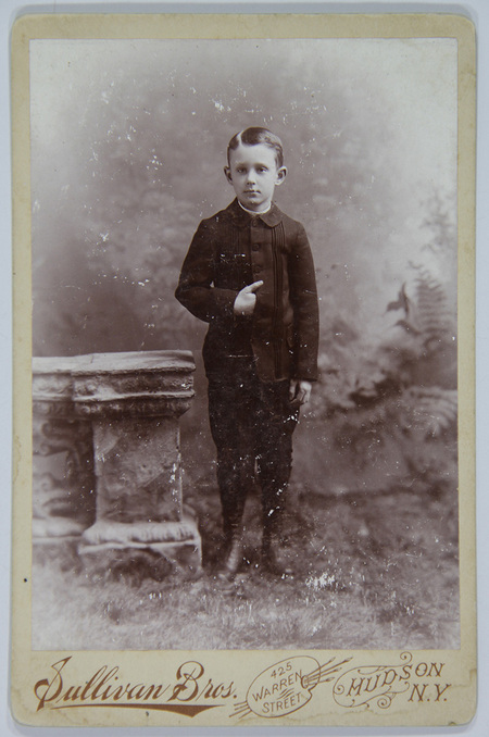 Retrato de estudio de un niño de pie junto a un pedestal de piedra ornamental. El menor luce un peinado formal con raya lateral y viste un traje oscuro con una chaqueta de diseño plisado en el frente, botones centrales y pantalones cortos tipo knickerbockers con medias oscuras. El fondo presenta un telón pintado con motivos de follaje brumoso.