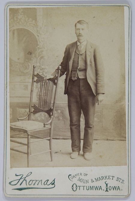 Retrato de estudio de un hombre de mediana edad con bigote, vestido con un traje formal de tres piezas y leontina de reloj visible en el chaleco. El sujeto posa de pie apoyando su mano sobre una silla de estilo victoriano con respaldo ornamentado, frente a un telón pintado con motivos clásicos.