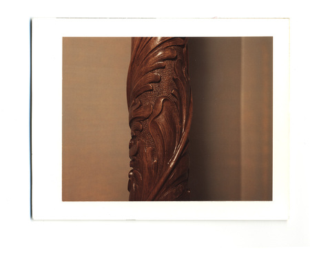 Detalle fotográfico de una columna o soporte de madera tallada con motivos ornamentales. La pieza presenta relieves profundos de hojas entrelazadas sobre una superficie con textura granulada, representativa de la artesanía en madera en contextos arquitectónicos o de mobiliario histórico mexicano, resaltada por una iluminación de estudio que enfatiza el volumen.