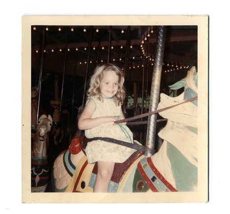 Una niña de cabello claro y rizado sonríe mientras monta un caballo de carrusel en lo que parece ser una feria o parque de atracciones. La escena captura un momento de ocio infantil característico de la fotografía familiar de mediados del siglo XX, destacando los detalles ornamentales del carrusel y las luces decorativas al fondo que evocan la atmósfera recreativa de la época.