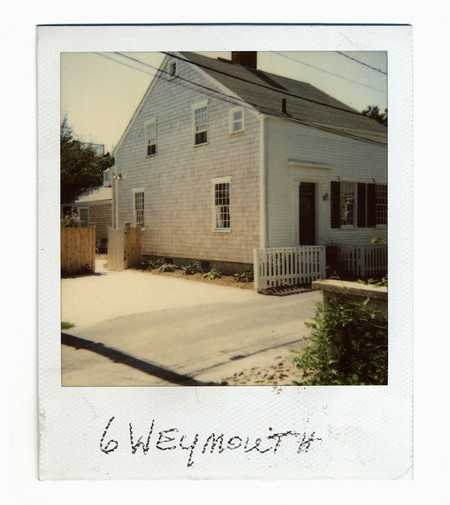 Fotografía a color de una residencia de estilo colonial tradicional de Nueva Inglaterra, identificada por la inscripción manuscrita como el número 6 de la calle Weymouth. La imagen muestra una casa con revestimiento de tejas de cedro y fachada lateral blanca, una cerca de estacas de madera y un muro de piedra en primer plano, capturando la estética residencial típica de la costa noreste estadounidense en la segunda mitad del siglo XX.