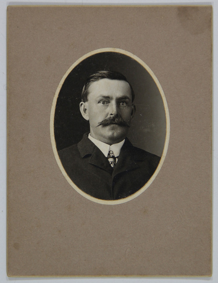 Retrato de estudio de un hombre de mediana edad con un bigote espeso y bien definido. Viste ropa formal de principios del siglo XX, que incluye un saco oscuro, camisa blanca de cuello alto rígido y una corbata con patrón gráfico. La fotografía está contenida en un marco ovalado impreso, montada sobre un soporte de cartón grueso de tono neutro que presenta un sello seco de estudio en la esquina inferior derecha.