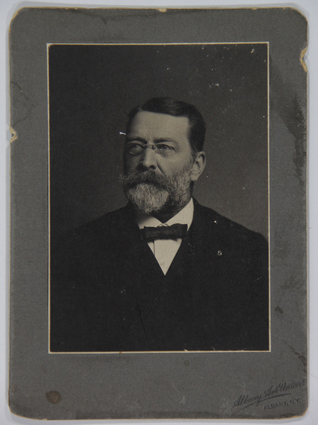 Retrato de estudio de un hombre de edad madura con anteojos redondos, barba densa y bigote entrecano. Viste indumentaria formal compuesta por una chaqueta oscura, camisa blanca de cuello rígido y una corbata de lazo (bow tie) oscura. El sujeto posa de tres cuartos mirando hacia un punto fuera de cámara.