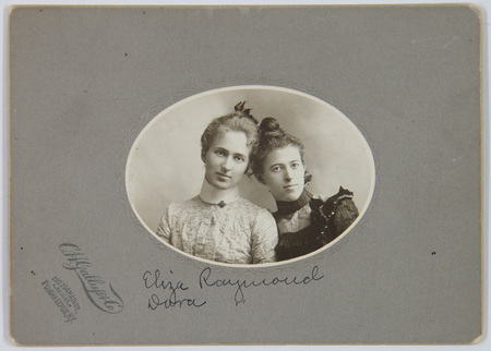Retrato de estudio de busto de dos mujeres jóvenes. Ambas visten indumentaria formal de la era victoriana tardía, con blusas de cuello alto y detalles ornamentales en encaje y seda. La imagen está centrada en un formato ovalado sobre un soporte de cartón que incluye el sello del fotógrafo y una identificación manuscrita de los sujetos.