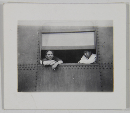 Retrato de dos hombres asomados por la ventana de un vagón ferroviario construido con placas de metal remachadas. El hombre de la izquierda viste una camisa oscura y observa directamente a la lente con los brazos apoyados en el marco, mientras que el hombre a su derecha, de complexión más robusta y camisa clara, mantiene la mirada baja en una actitud reflexiva durante lo que parece ser una parada del trayecto.