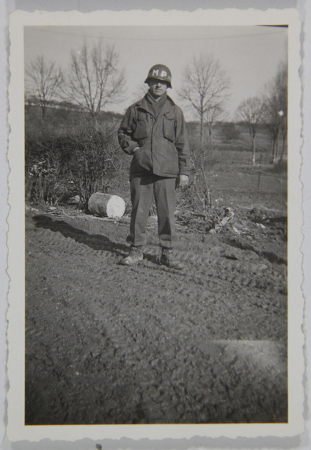 Retrato de cuerpo completo de un hombre joven uniformado como Policía Militar (MP). El sujeto posa de pie frente a un paisaje rural invernal con árboles desnudos y un suelo lodoso que muestra huellas de neumáticos de gran tamaño. Viste una chaqueta de campo M-1943, pantalones de lana y casco reglamentario marcado, situando la escena probablemente en Europa a mediados de la década de 1940.