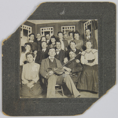 Un retrato grupal de quince individuos, hombres y mujeres, posando de manera formal en un entorno interior decorado con paneles acristalados. El foco central es un hombre sentado que sostiene un violín. Los integrantes presentan una vestimenta típica de la clase media o alta de principios del siglo XX, sugiriendo un evento social o familiar.