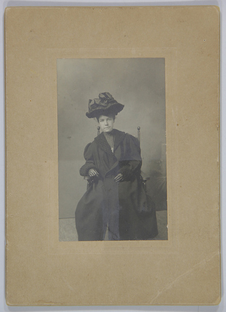 Retrato de estudio de una mujer joven en posición sedente, capturada con una pose formal. La protagonista viste una indumentaria invernal elegante compuesta por un abrigo oscuro de textura pesada con ribetes de piel en cuello y puños, complementado con un sombrero prominente adornado con grandes lazos de tela.