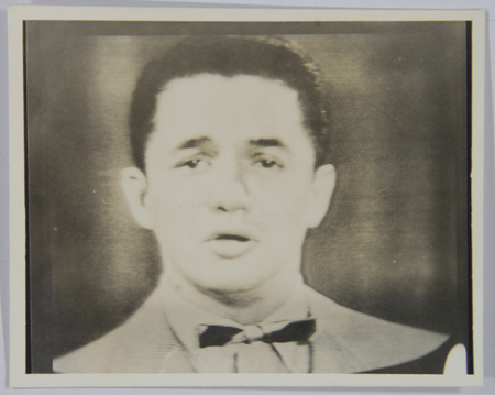 Retrato masculino de plano medio corto que muestra a un hombre joven con camisa clara y moño oscuro. El sujeto mira directamente a la camara con una expresion neutra y la boca ligeramente entreabierta. La imagen se caracteriza por una textura de lineas horizontales paralelas en toda su superficie y se encuentra enmarcada por un borde blanco de papel fotografico.