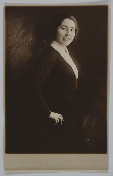 Retrato de estudio de una mujer joven capturada en una pose de tres cuartos. Viste una chaqueta oscura con un cuello claro prominente, caracteristico de la moda de las primeras decadas del siglo XX. Su expresion es afable, mirando directamente a la camara con una mano apoyada en la cadera sobre un fondo neutro texturizado.