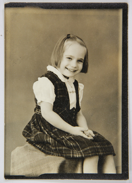 Retrato de estudio de una niña pequeña sentada, con una expresión sonriente. Viste un atuendo escolar o de gala compuesto por un vestido de cuadros oscuros y una blusa blanca de manga corta con bordados en el pecho. La imagen captura la estética de la infancia urbana en el México de mediados del siglo XX.