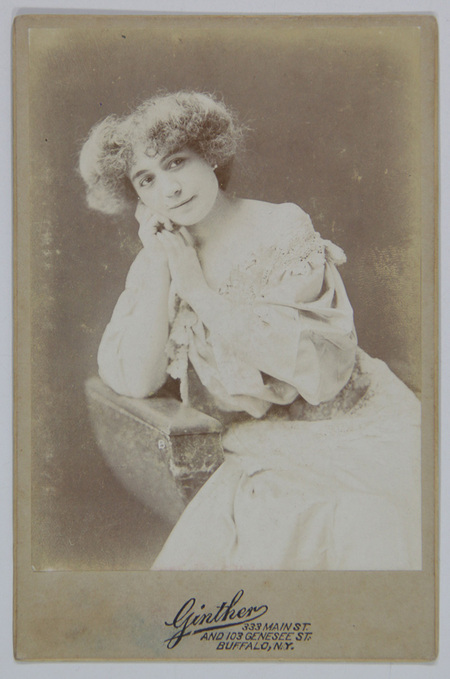 Retrato de estudio en plano medio de una mujer joven con cabello corto y rizado. La figura aparece apoyada sobre un mueble de utilería, sosteniendo su rostro con ambas manos en una pose introspectiva. Viste un vestido de tono claro con un prominente cuello de encaje, típico de la estética de finales de la época victoriana.
