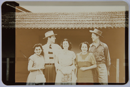 Un grupo de cinco personas, tres mujeres y dos hombres, posan sonrientes frente a una edificacion de estilo rural con techo de lamina. Los hombres visten sombreros y camisas de manga larga, destacando uno con una corbata de diseño audaz. Las mujeres lucen vestidos de corte mid-century, una de ellas porta un bolso blanco de mano y otra un vestido con patron de lunares. La disposicion sugiere una fotografia de convivencia social o familiar en un entorno campestre mexicano de mediados del siglo XX.