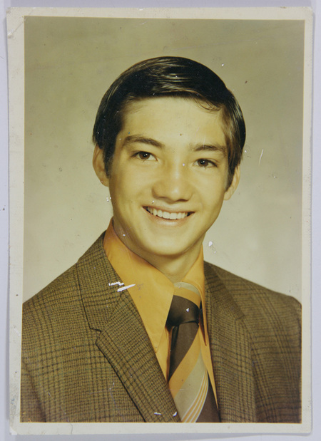 Retrato de estudio individual de un hombre joven en encuadre de busto. El sujeto posee rasgos mestizos, cabello oscuro peinado hacia un lado y expresión sonriente. Viste un atuendo formal compuesto por un saco con diseño de cuadros pequeños, camisa naranja brillante de cuello largo y corbata ancha con franjas diagonales en tonos tierra, típica de la moda urbana mexicana de los años 70.