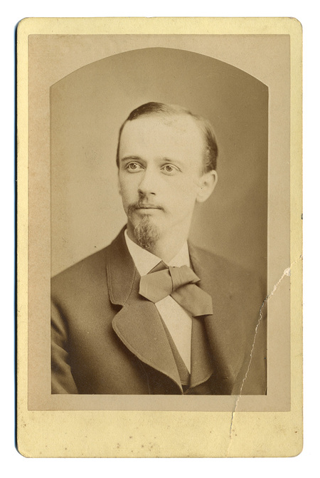 Retrato de estudio en plano medio de un hombre joven con mirada dirigida hacia su derecha. El sujeto viste un traje formal oscuro de solapas anchas, camisa de cuello rígido y un corbatín de lazo prominente. Su fisonomía destaca por una frente amplia, bigote delgado y una barba de tipo perilla cuidadosamente recortada.