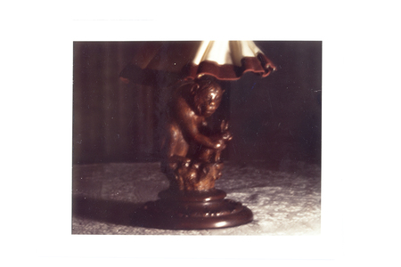 La imagen muestra una lámpara de mesa decorativa situada sobre una superficie cubierta por un mantel de encaje blanco. La base de la lámpara consiste en una figura tallada que representa a un primate en un tono marrón oscuro. La pantalla es de color crema con bordes ondulados en color marrón rojizo, capturada en un ambiente interior con iluminación artificial tenue.