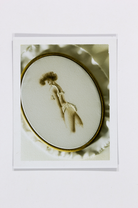 Fotografía instantánea que registra una pieza de arte textil. Se observa un bastidor ovalado con un bordado que representa la silueta de una mujer desde una perspectiva posterior, vistiendo lencería blanca. La figura está realizada en tonos ocres y sepias, contrastando con la blancura de las prendas bordadas y el fondo de tela fruncida que sirve como escenario.