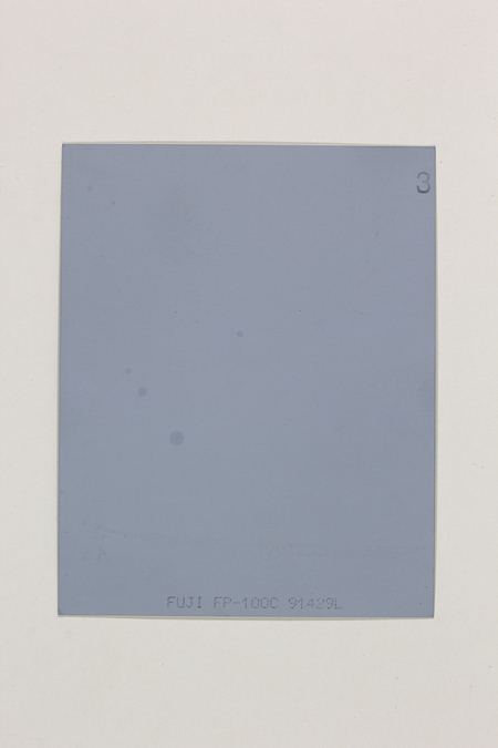 La imagen muestra el reverso o una hoja descartada de una película instantánea Fuji FP-100C. Presenta una superficie de tono gris azulado uniforme con marcas técnicas de fabricación en la parte inferior y un número tres manuscrito en la esquina superior derecha. No contiene elementos figurativos ni escena fotográfica capturada.