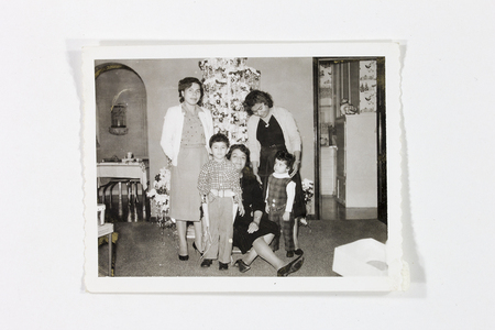 Retrato grupal de una familia en un interior doméstico durante una celebración navideña. Tres mujeres de distintas edades y dos niños pequeños posan frente a un árbol de Navidad decorado con festones y esferas. La escena muestra una estancia con un arco de medio punto y una vista hacia una cocina con papel tapiz decorativo, capturando la estética hogareña mexicana de mediados del siglo XX.