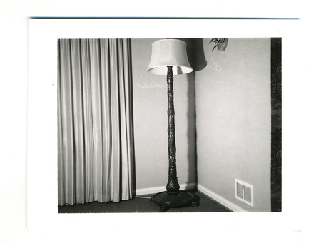 Una fotografía de un rincón interior que destaca una lámpara de pie ornamentada con fuste tallado y una pantalla de tela. A la izquierda se observan cortinas de pliegues verticales y a la derecha una pared con una rejilla de ventilación cerca del suelo, reflejando el diseño decorativo de mediados del siglo XX.