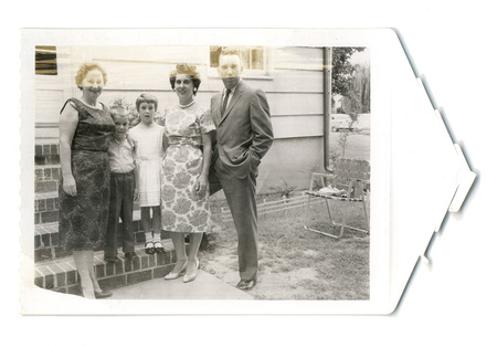 Fotografía grupal de una familia compuesta por dos mujeres, un hombre y dos niños posando frente a una vivienda. Las mujeres visten elegantes vestidos de época y perlas, mientras el hombre porta un traje formal y los niños ropa dominical. Se encuentran junto a una escalinata de ladrillo en un entorno de jardín doméstico que incluye una silla de patio y un automóvil visible al fondo, capturando un momento de la vida social de mediados del siglo XX.