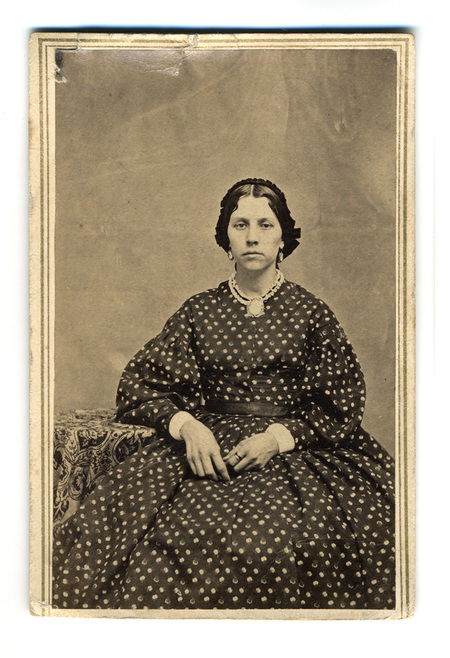 Retrato de estudio de una mujer joven de mediados del siglo XIX. La sujeta aparece sentada, vistiendo un vestido oscuro con patron de puntos, mangas amplias y una falda voluminosa. Porta un tocado oscuro, aretes y un collar con un camafeo central. A su lado se observa una mesa cubierta con un tapete de motivos fitomorfos, tipico de la utileria de estudio de la epoca.