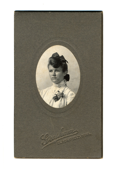 Retrato de estudio de una joven mujer en plano medio corto, presentado en un óvalo. Viste una blusa clara de cuello alto con encajes, un broche central y un ramillete floral. Su cabello está recogido con un lazo oscuro de gran tamaño, característico de la estética de principios del siglo XX.