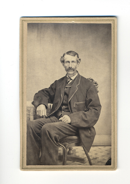 Retrato de estudio de un hombre de mediana edad con bigote y cabello ondulado, posando sentado. Viste un traje formal de tres piezas con un saco tipo 'sack coat' y chaleco. Se encuentra en un entorno controlado con una silla de madera tallada y una mesa con mantel decorado, elementos típicos de la escenografía fotográfica decimonónica.