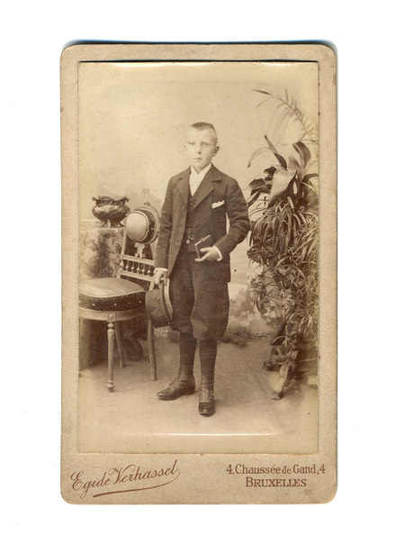 Retrato de estudio de un niño de pie, vestido con un traje formal oscuro de tres piezas: chaqueta, chaleco y pantalones tipo bombacho. El sujeto sostiene un sombrero en su mano derecha y un pequeño objeto rectangular, posiblemente un libro, en la izquierda. El entorno incluye una silla decorativa, una mesa con mantel floral y plantas ornamentales de gran tamaño al fondo.