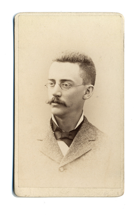Retrato de estudio de un hombre joven captado de tres cuartos. El sujeto posee cabello corto, un bigote denso y utiliza anteojos circulares. Viste una chaqueta de textura gruesa con solapas pequeñas, una camisa de cuello alto y un corbatín oscuro. La composición y el fondo neutro son típicos de la retratística comercial decimonónica.