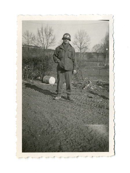 Retrato de cuerpo entero de un soldado de la Policía Militar estadounidense en un entorno rural lodoso. El individuo viste una chaqueta de campo M-1943, casco con las siglas 'MP' y botas de combate. La escena, capturada en exteriores con árboles caducifolios al fondo, evoca el periodo de la Segunda Guerra Mundial o la posguerra inmediata.