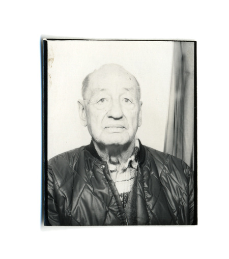Retrato masculino de plano medio corto donde aparece un hombre de edad avanzada con expresion serena. El sujeto viste una chaqueta acolchada de color oscuro sobre una camisa de franela a cuadros y un chaleco. La fotografia presenta una iluminacion directa y frontal con un fondo claro donde se aprecia ligeramente la caida de una cortina en el lado derecho.
