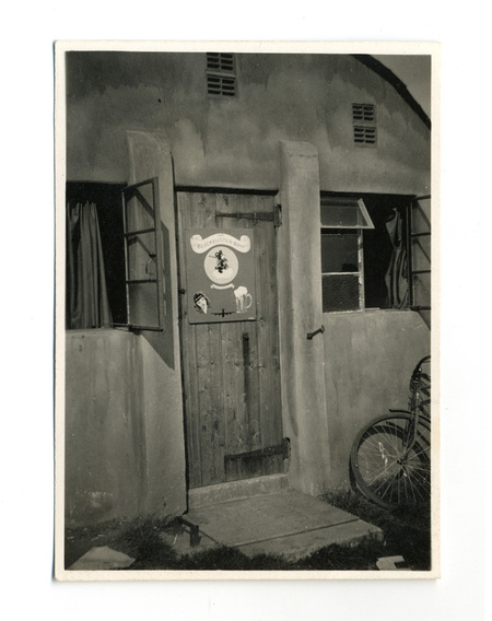 Vista exterior de la entrada a un alojamiento militar, probablemente un cuartel de tripulación aérea durante la Segunda Guerra Mundial. En la puerta de madera destaca un cartel titulado 'The Blockbuster Boys', ilustrado con arte gráfico que incluye el rostro de una mujer, un bombardero y una cerveza. Una bicicleta se encuentra apoyada contra la pared lateral.