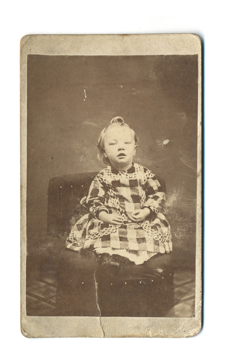 Retrato de estudio de un infante de corta edad, sentado sobre un sillón tapizado con flecos decorativos. El niño o niña viste una prenda a cuadros de gran formato con detalles de encaje o pasamanería en el cuello y las mangas. La pose es estática y formal, característica de la fotografía comercial del siglo XIX.